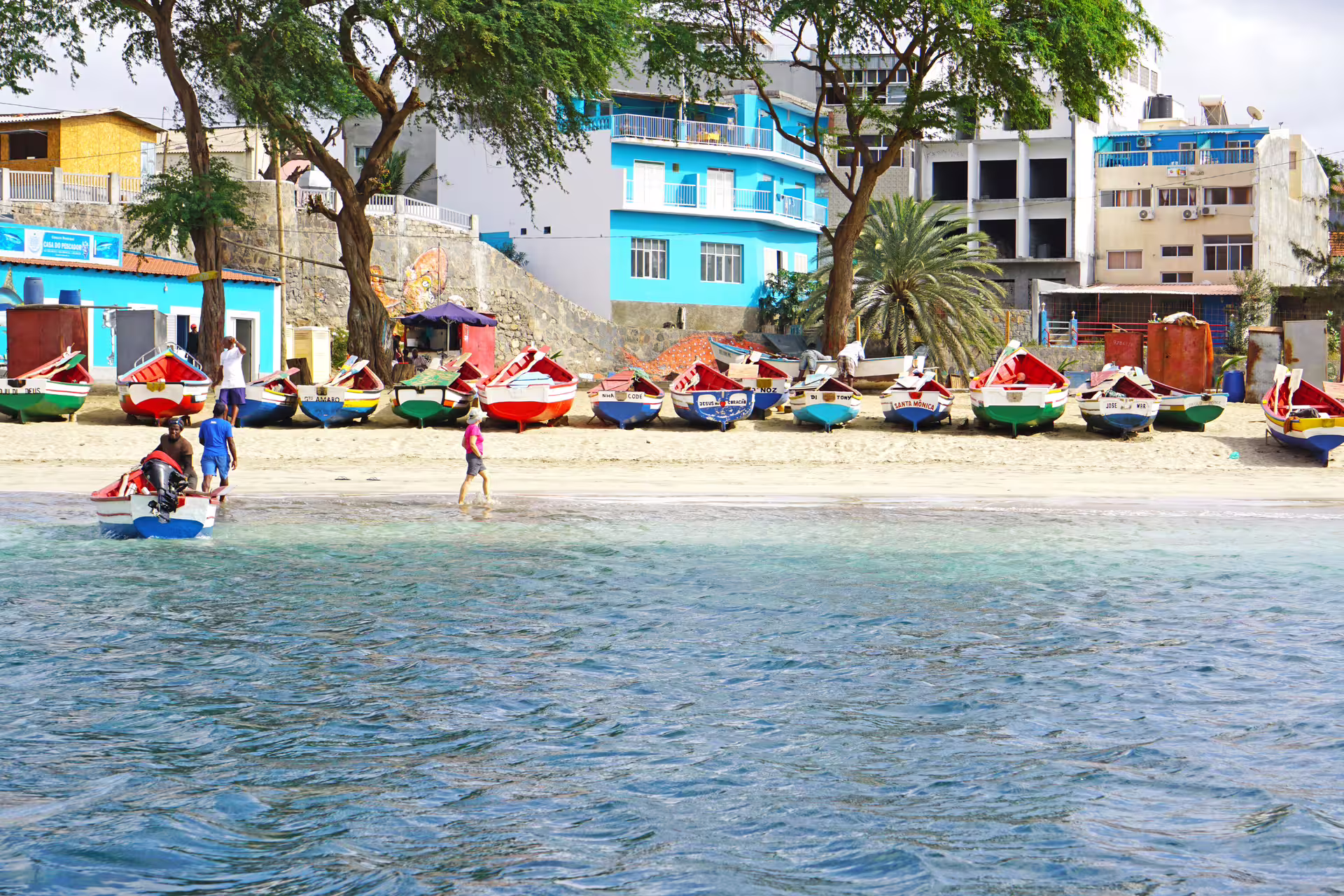 Colorful boats line the sandy beach near vibrant buildings at Tarrafal, with clear blue water in the foreground.
