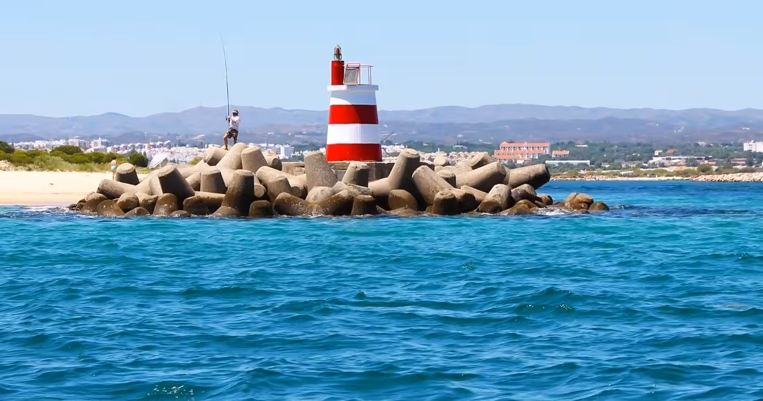 Red and white striped lighthouse on rocky breakwater with fisherman during Tavira Ria Formosa boat tour