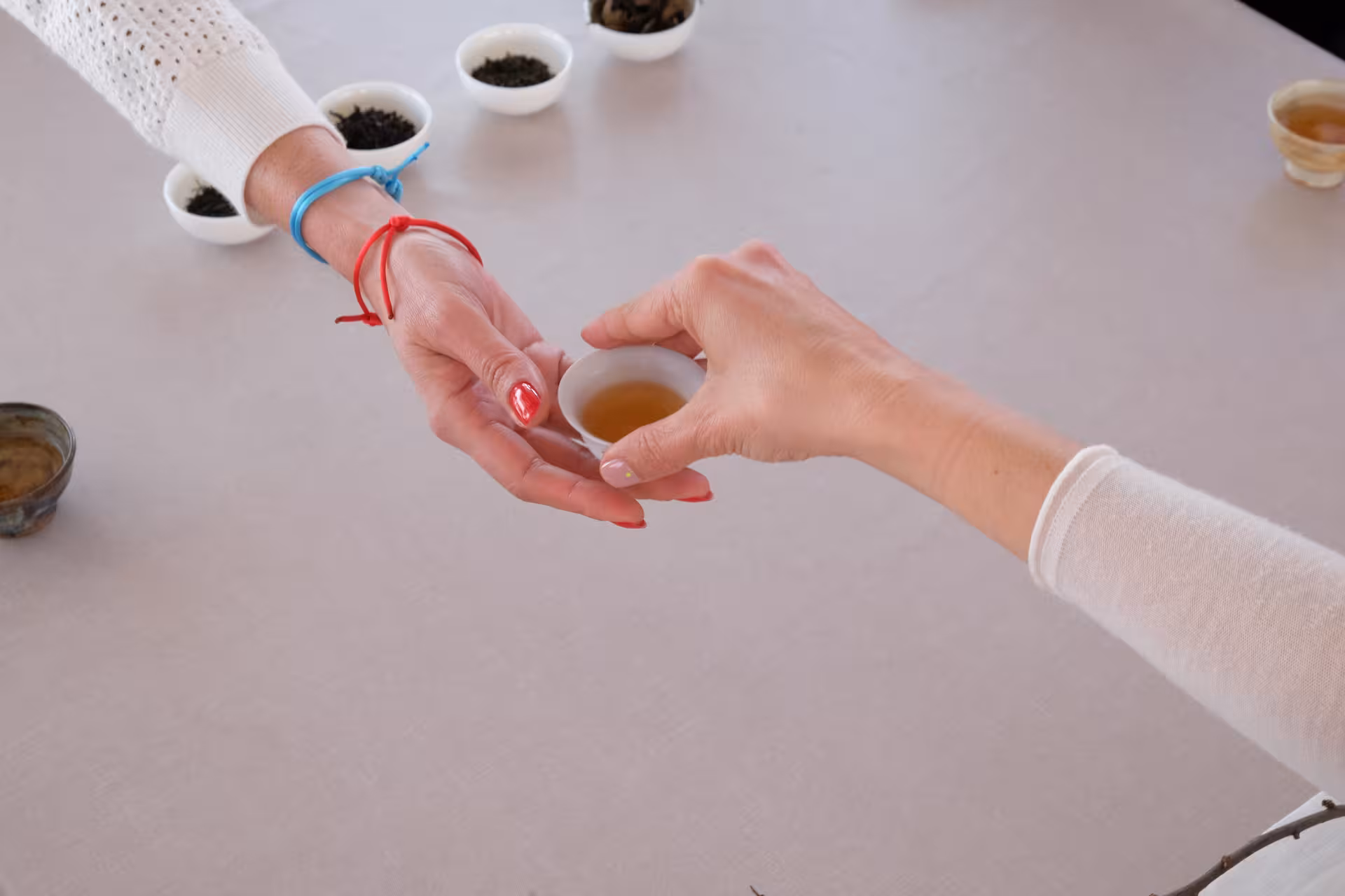 Hands pass a small cup of brewed tea during tea ceremony tasting, with loose leaf tea bowls in background