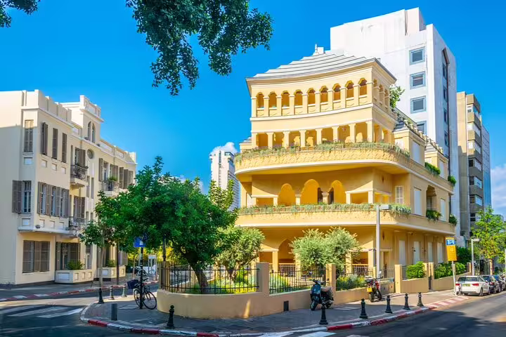 Yellow Bauhaus building on a sunny Tel Aviv street corner with lush trees and balconies