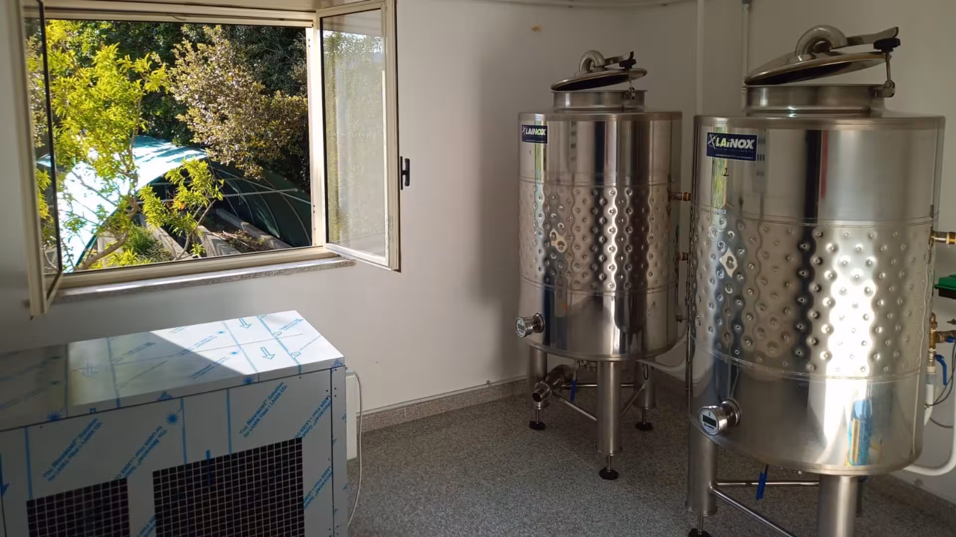 Stainless steel fermentation tanks in a sunlit room at the Tempio Pausania distillates workshop with garden view.