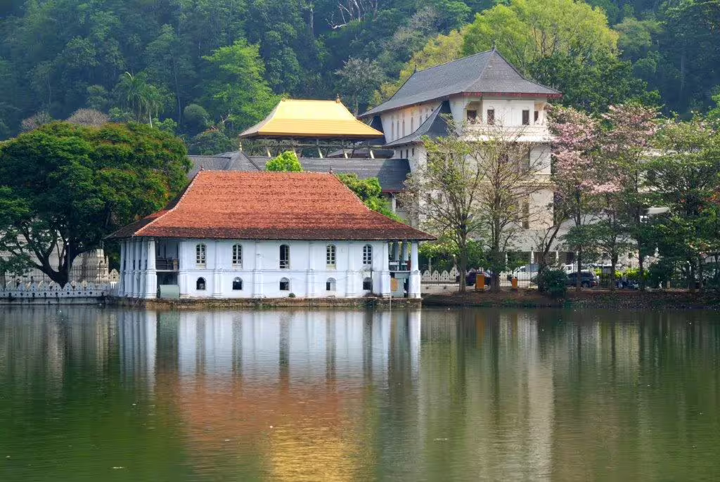 Serene view of the Temple of the Sacred Tooth Relic in Kandy, Sri Lanka, reflecting in the calm lake surrounded by lush greenery.