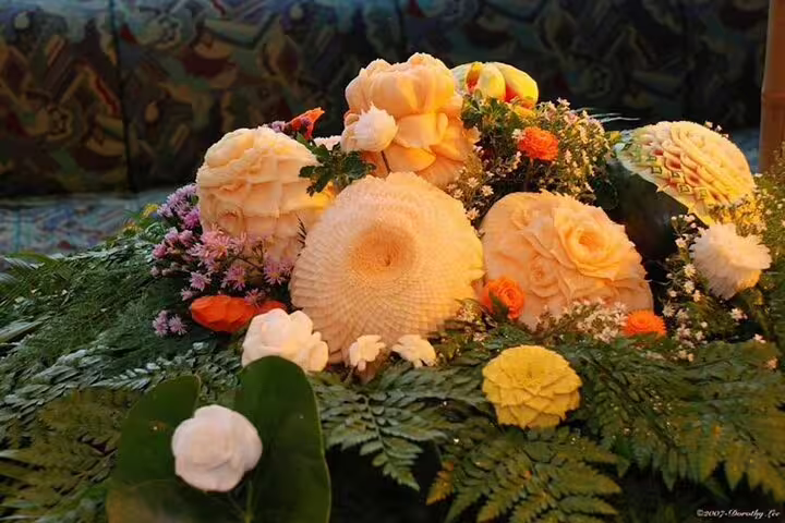 Elegant Thai fruit carving display featuring intricate floral designs on melons surrounded by fresh greenery and flowers