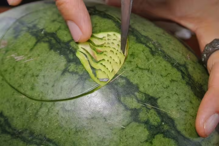 Hands skillfully carving decorative patterns into a watermelon during a Thai fruit carving class