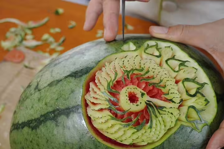 Skilled hands carving an intricate floral dragon design into a fresh watermelon during a Thai fruit carving class