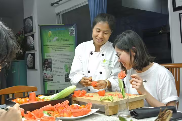 Smiling Thai chef instructor and student enjoying a hands-on tomato flower carving lesson in a Thai carving class