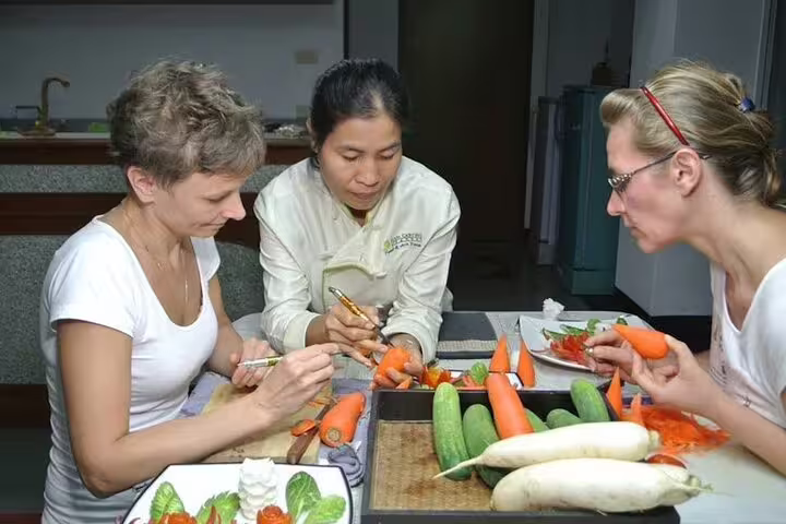 Thai chef guides two students in traditional fruit and vegetable carving class using carrots and cucumbers