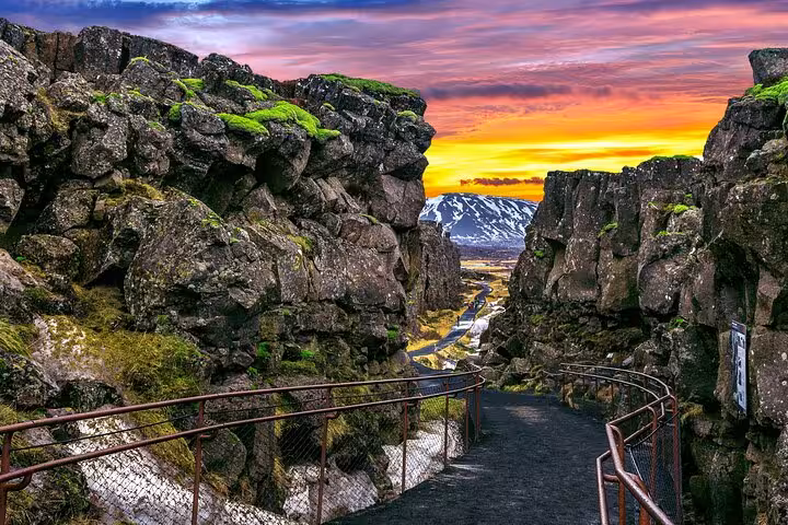 Narrow rocky canyon path with moss-covered walls at golden sunset, snowy mountain in background, Þingvellir Iceland