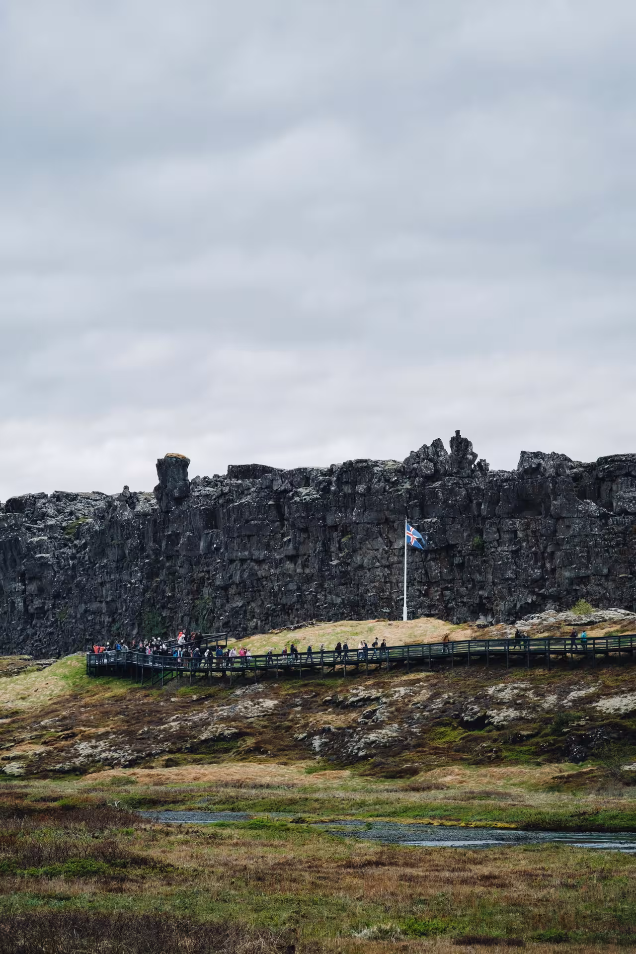 Tourists walking boardwalk at Þingvellir National Park with Icelandic flag and dramatic lava rock cliffs