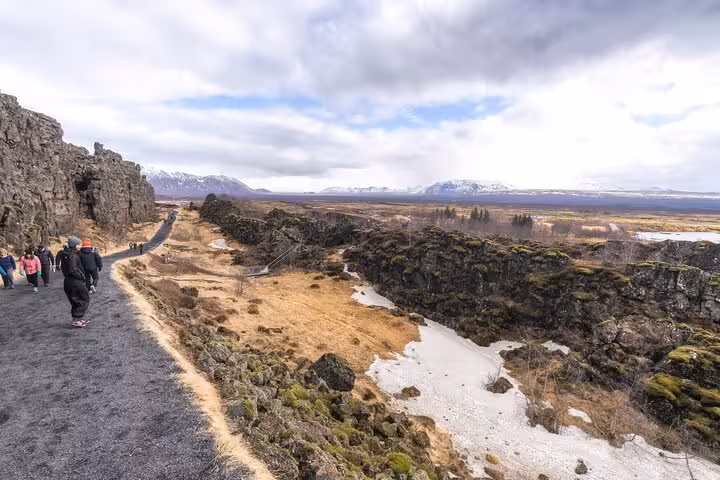 Tourists walking along rocky Þingvellir rift valley path with snow-capped mountains on Golden Circle tour