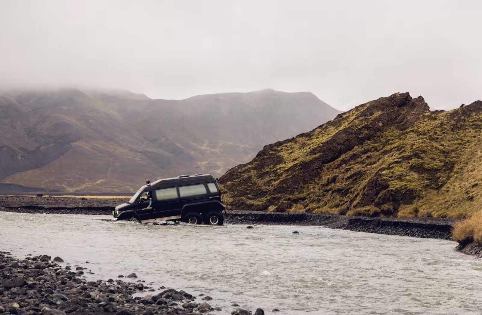 Black 6x6 off-road van crossing glacial river in Thorsmork Iceland with misty mountains