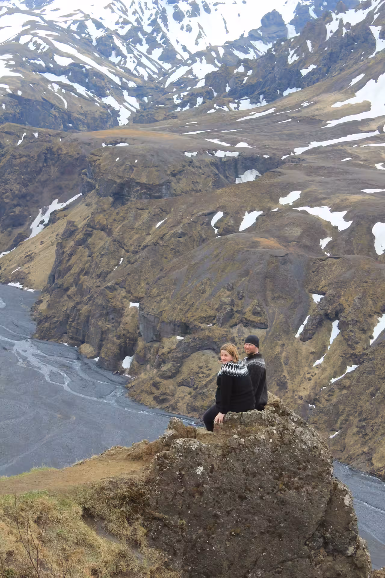 Couple in Icelandic sweaters sitting on rocky outcrop overlooking Thorsmork canyon and snowy mountains