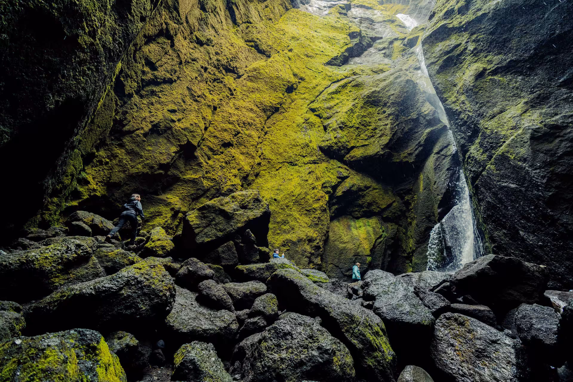 Hikers climbing mossy rocks inside a dramatic narrow canyon with a waterfall in Thorsmork Iceland