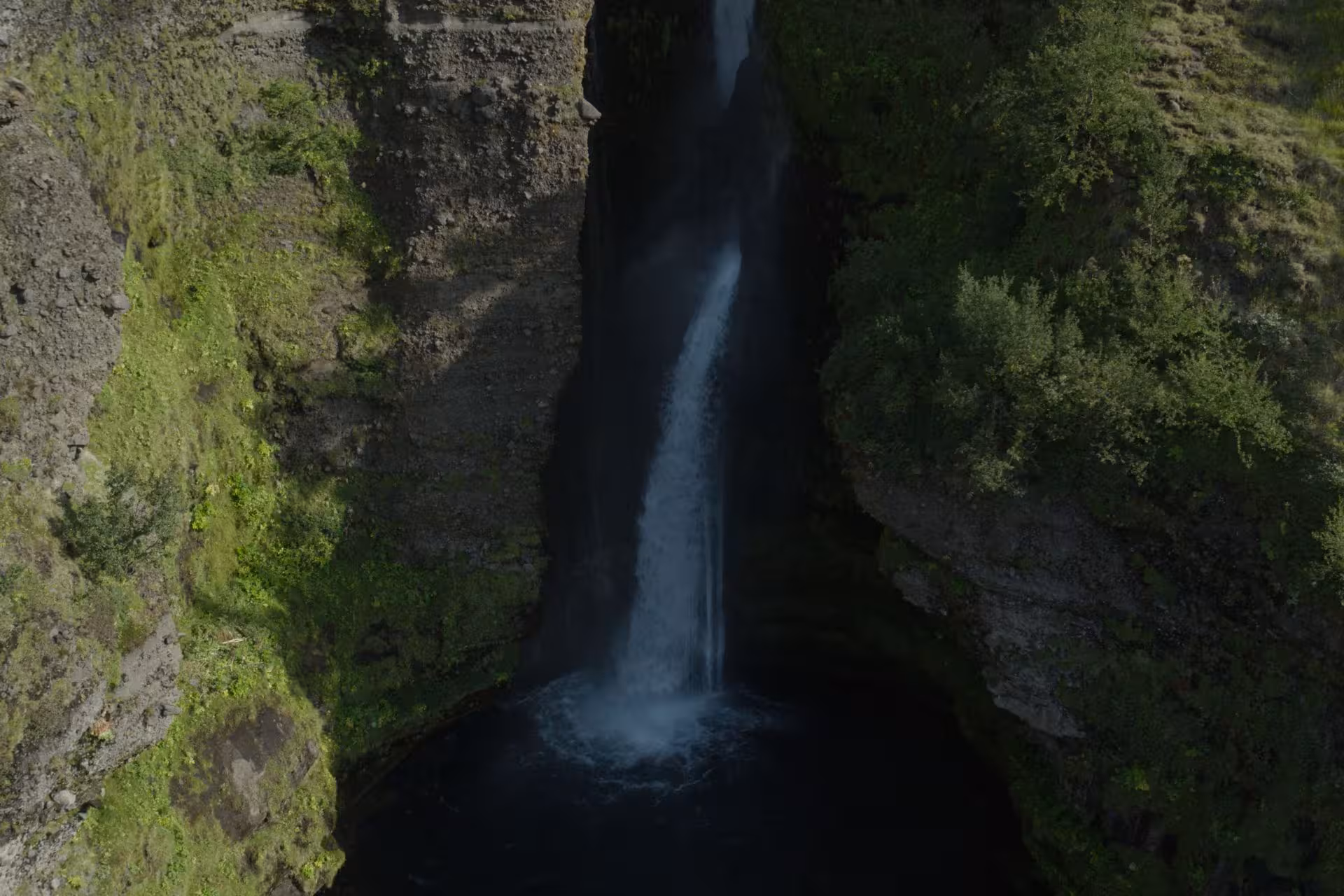 Narrow canyon waterfall plunging into dark pool surrounded by mossy volcanic cliffs in Thorsmork highlands
