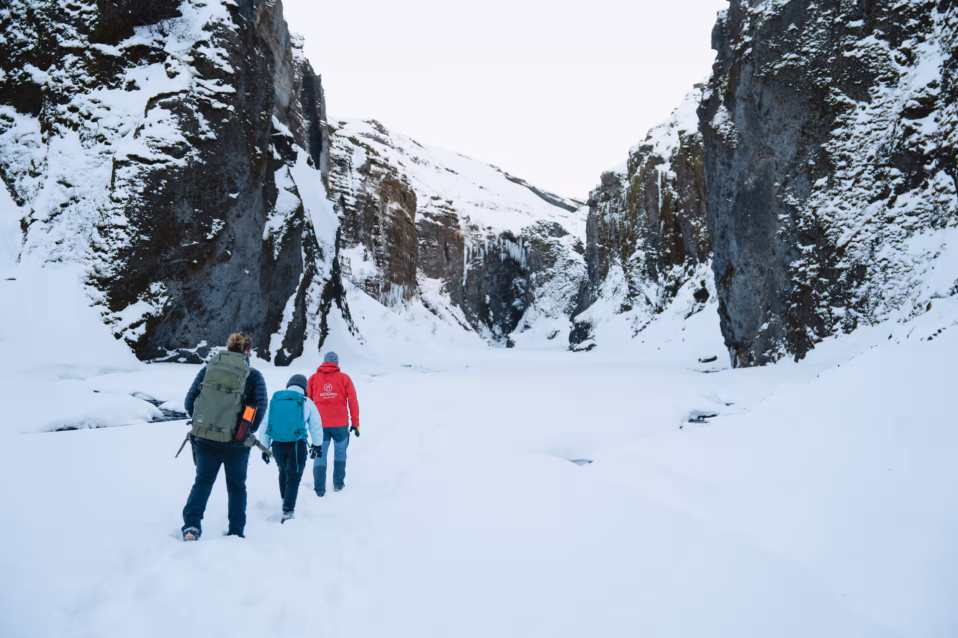 Three hikers walking through snow-covered Thorsmork canyon with towering rocky cliffs in winter, Iceland