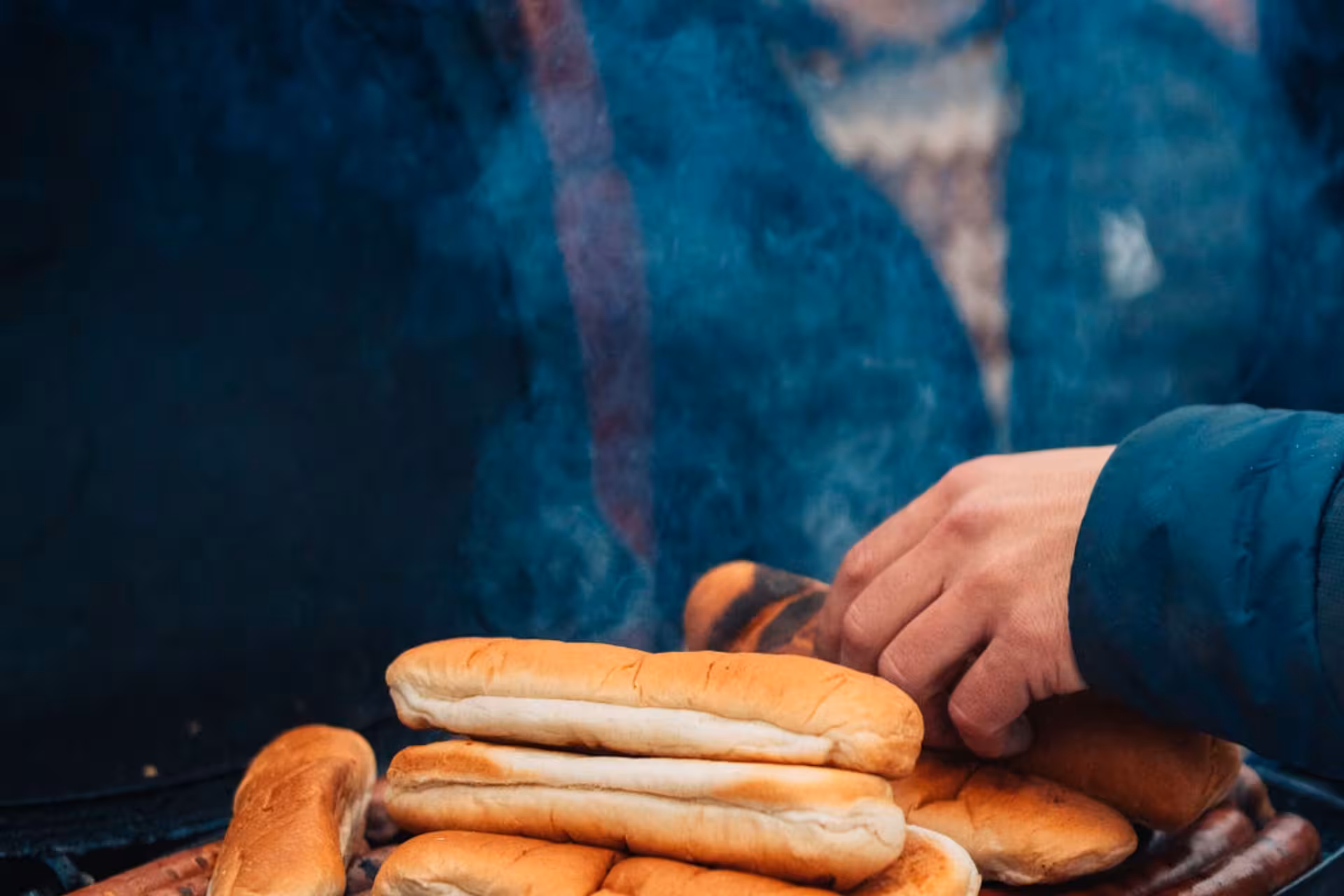 Hand placing hot dog buns on a smoky grill with sausages during an outdoor tour