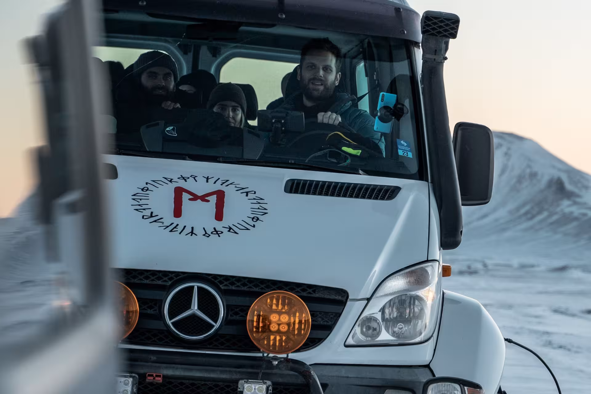 Three passengers in a white Mercedes truck with runic logo driving through snowy Icelandic highlands