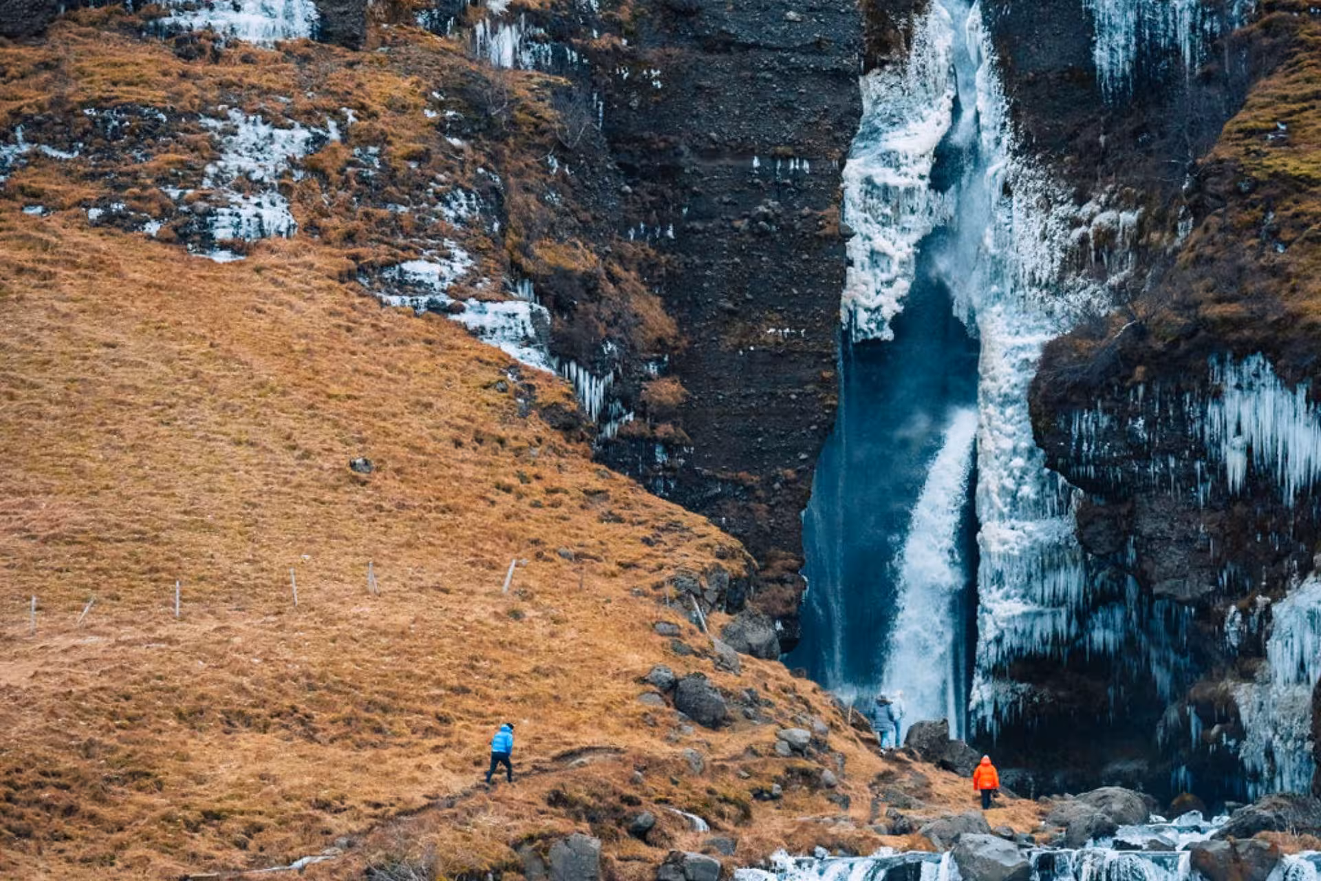 Two hikers in colorful jackets approaching a partially frozen waterfall in Iceland's wild highlands