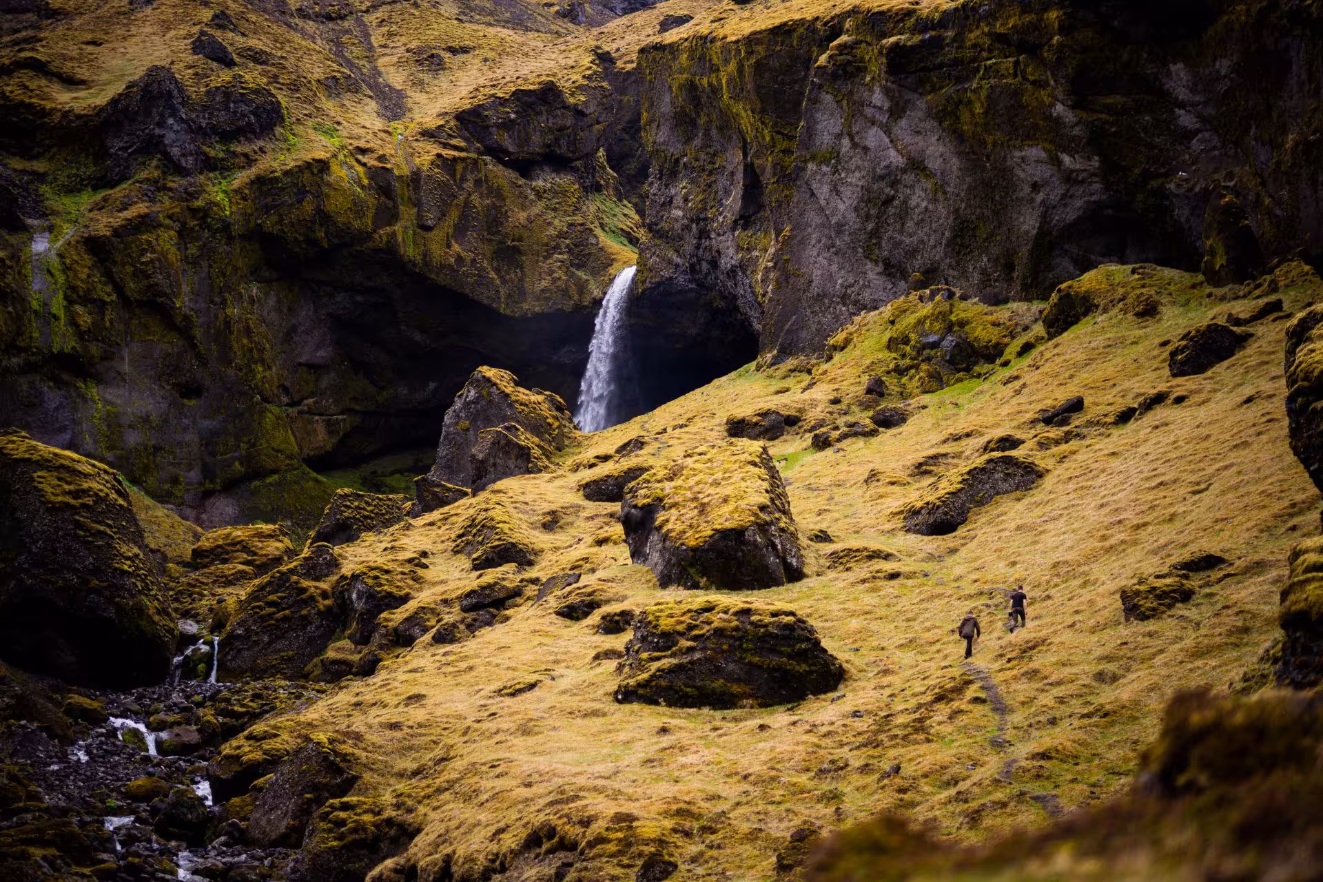 Two hikers trekking toward a hidden waterfall in a mossy lava canyon in Thorsmork, Iceland