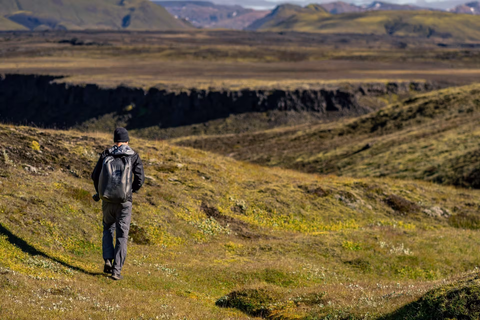 Hiker with backpack walking through green mossy highlands toward volcanic canyon in Thorsmork, Iceland
