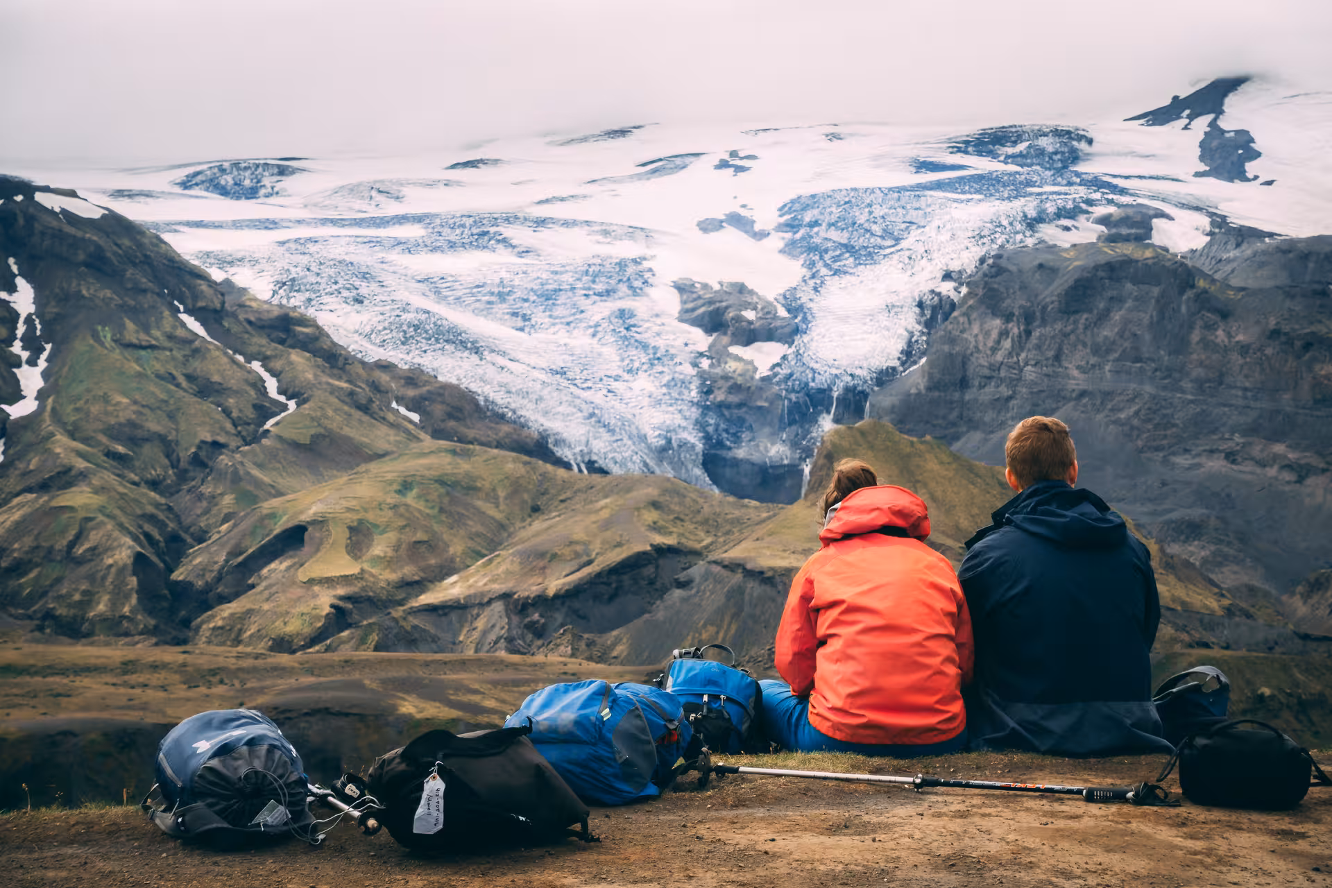 Two hikers resting with backpacks overlooking a glacier and green volcanic mountains in Iceland