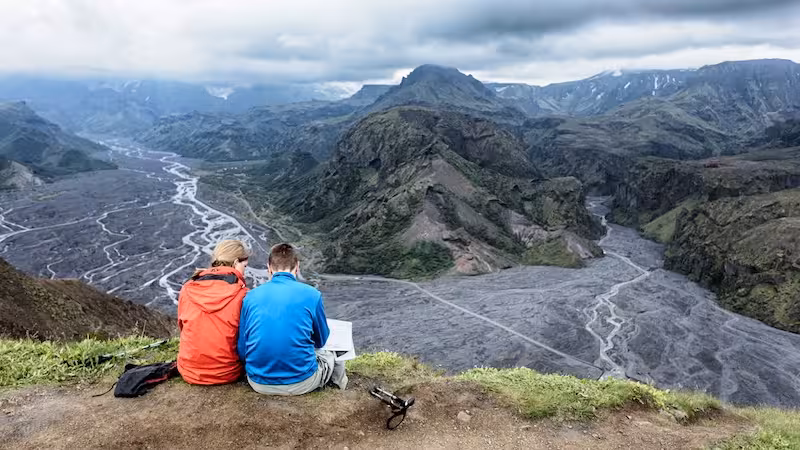Two hikers sitting on a cliff edge overlooking Thorsmork valley with braided rivers and glaciers in Iceland