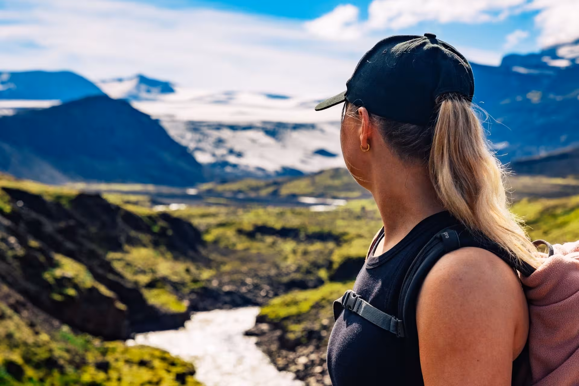 Hiker with blonde ponytail and black cap gazing at glacier valley and river in Thorsmork, Iceland