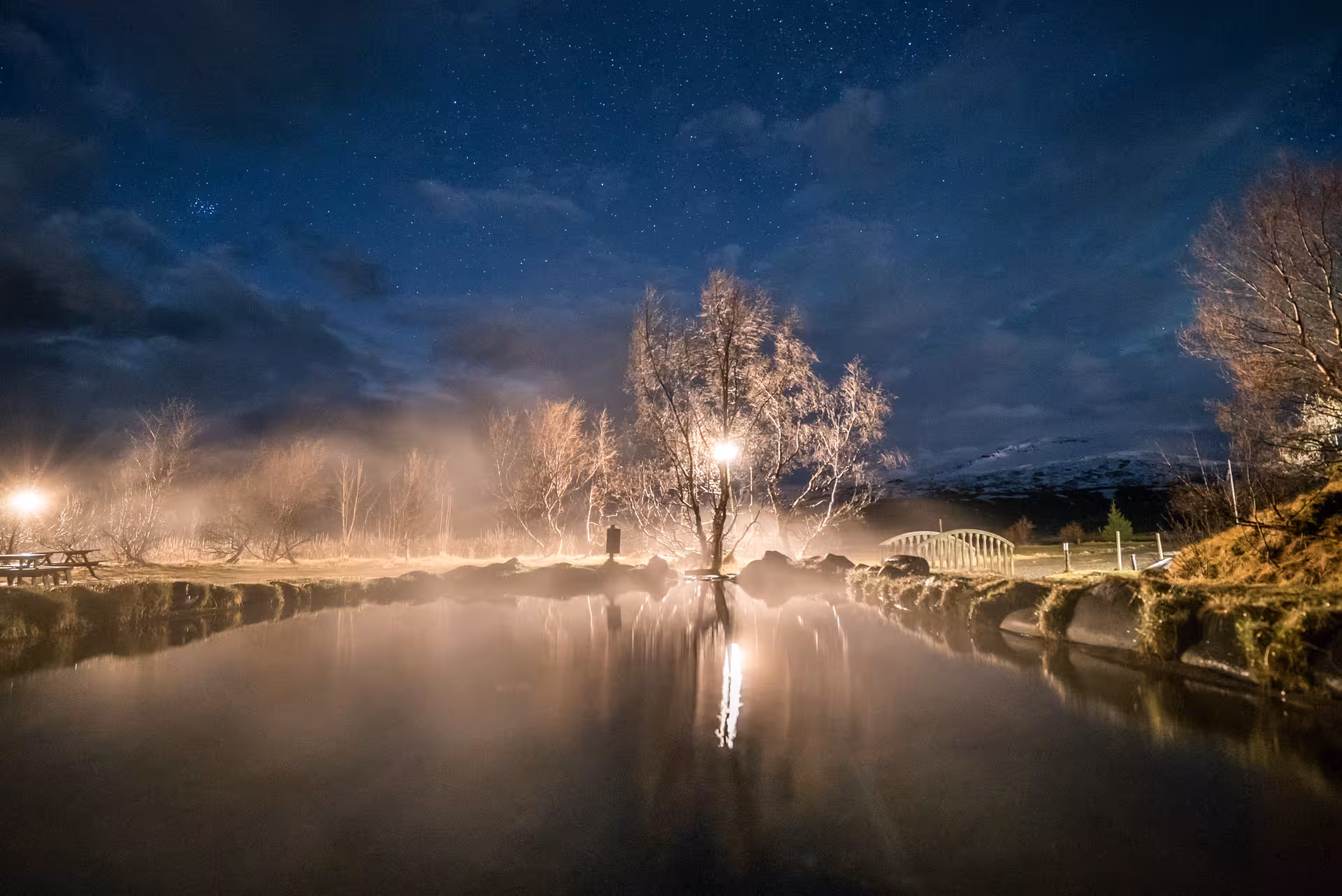 Misty geothermal hot spring pool at night with starry sky and glowing street lights reflecting on water, Iceland