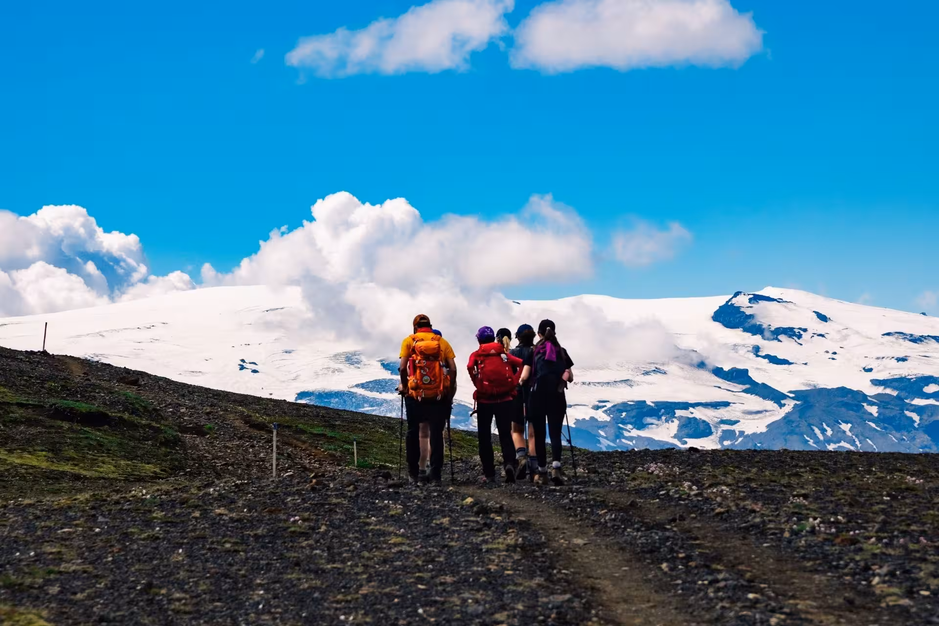 Four hikers with backpacks and trekking poles on a rocky trail heading toward snow-capped mountains in Thorsmork, Iceland