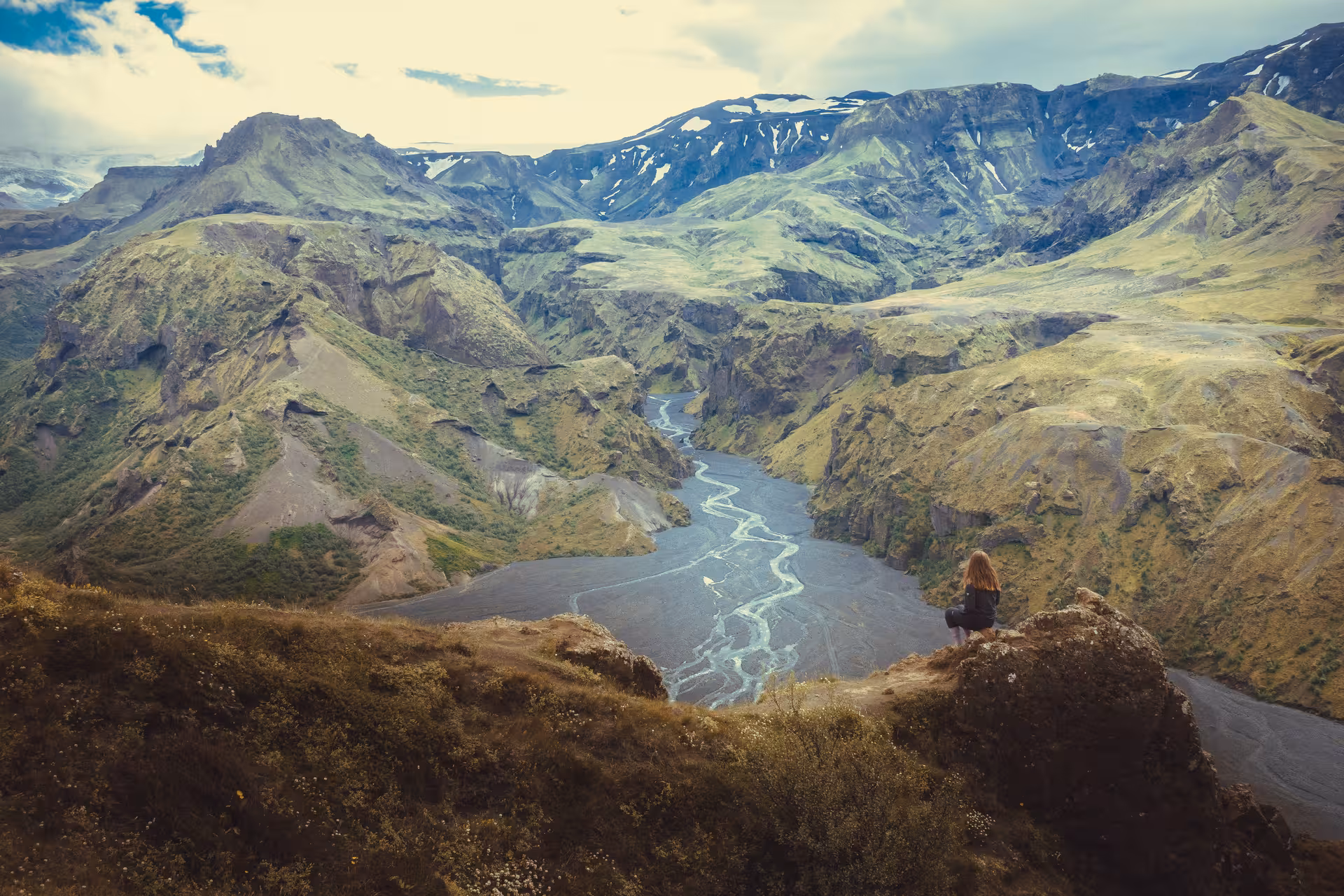 Hiker sitting on cliff overlooking Thorsmork canyon river valley with green mountains and snow patches