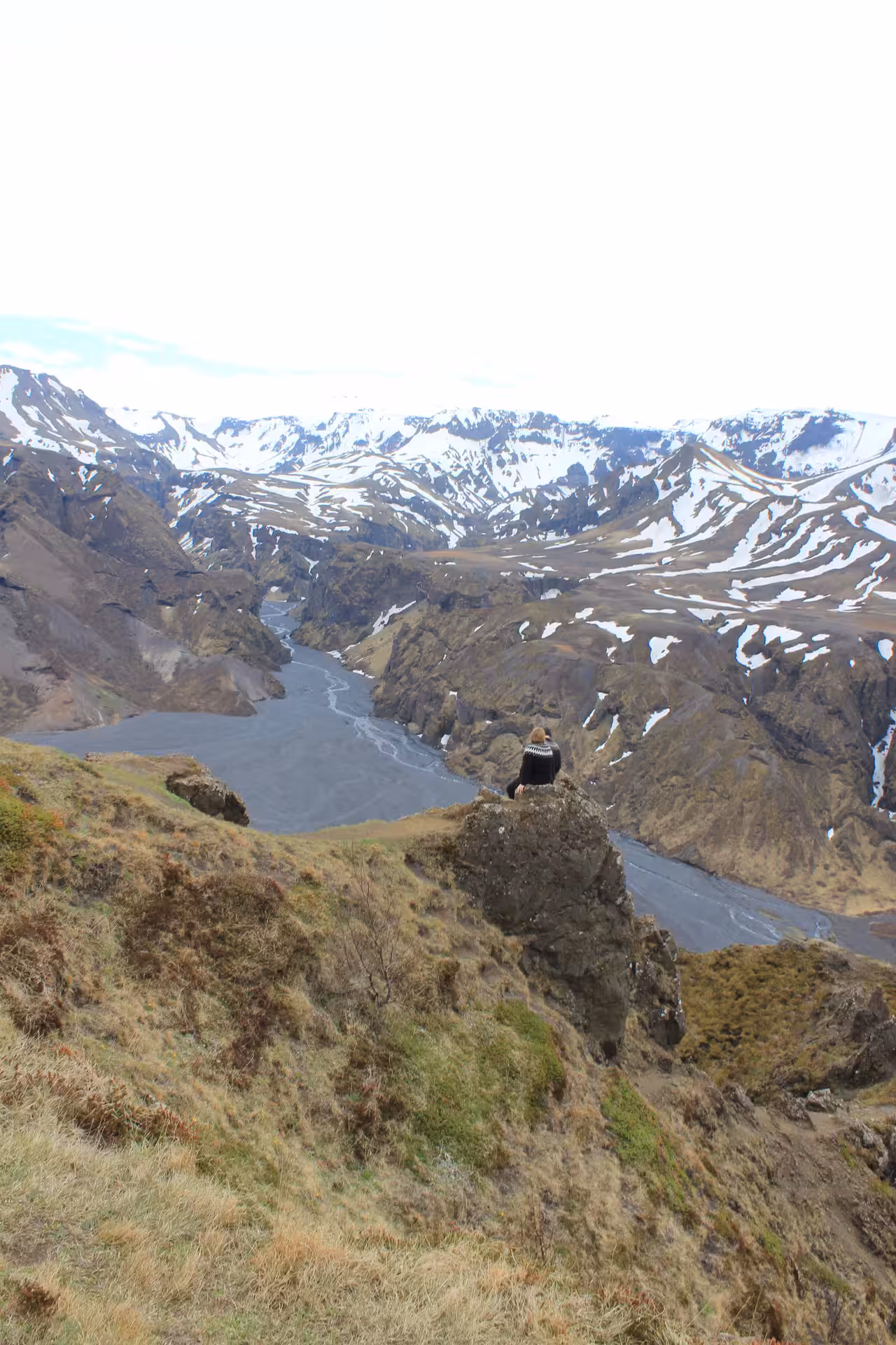 Hiker sitting on rocky outcrop overlooking Thorsmork river canyon and snow-capped mountains in Iceland