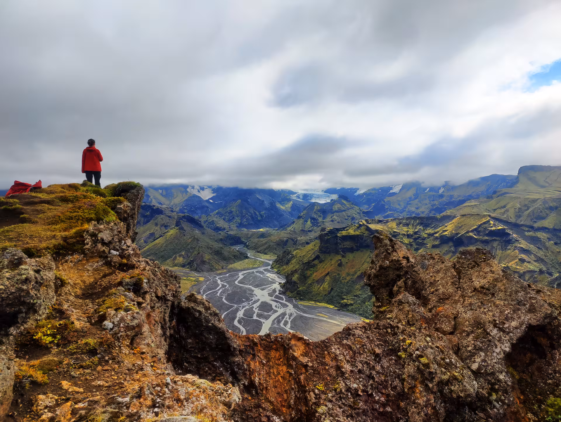 Hiker in red jacket standing on cliff overlooking Thorsmork valley with braided river and glacier in South Iceland