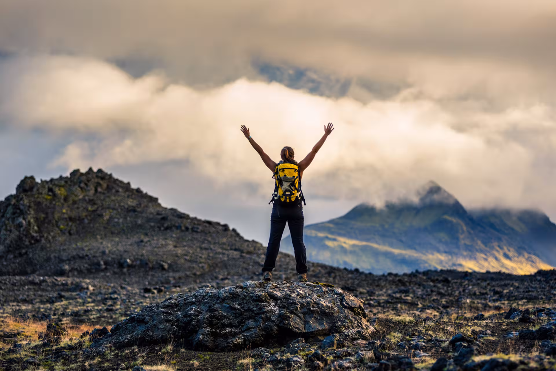 Hiker with arms raised on volcanic rock with dramatic clouds over South Iceland mountains