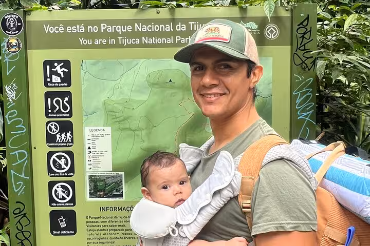 Smiling visitor with baby in front of Tijuca National Park sign, ready for an adventure in the lush forest.