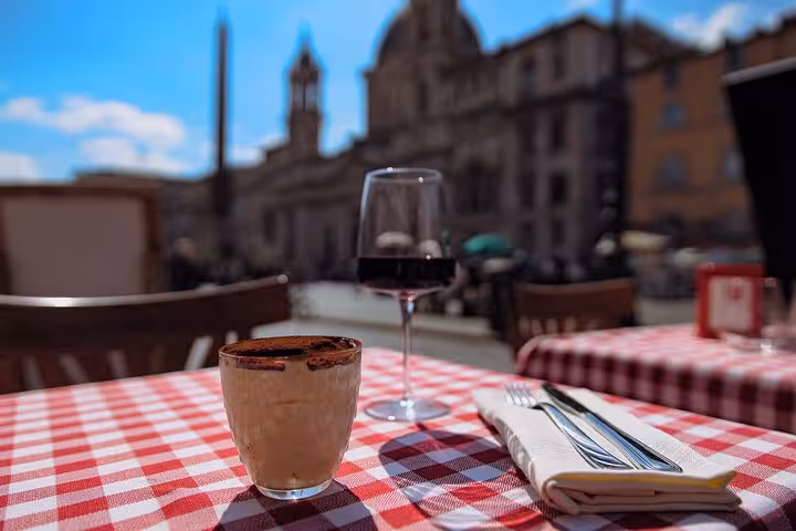 Tiramisu and red wine on a checkered tablecloth with Rome's Piazza Navona in the background.