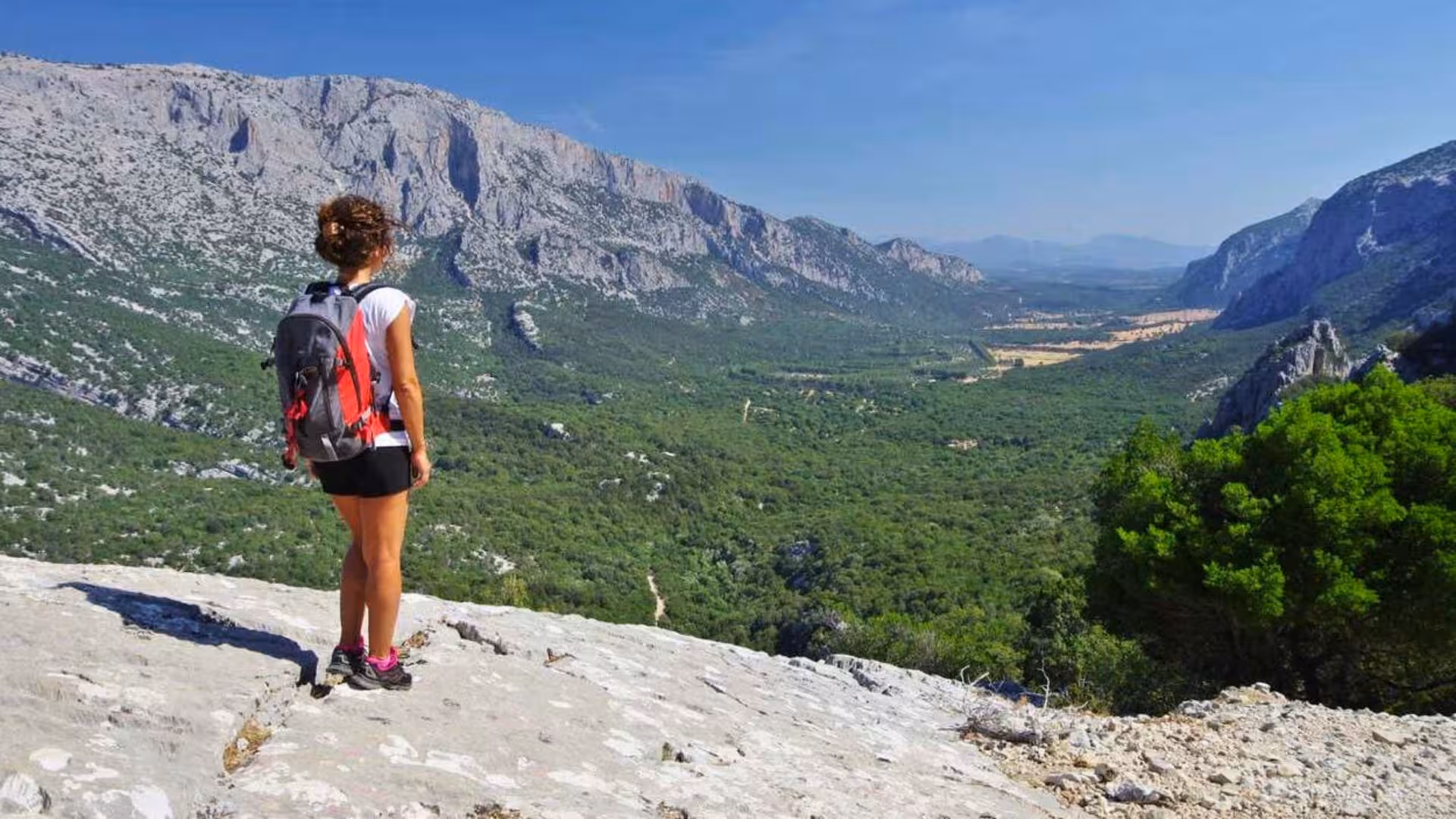 Hiker enjoys panoramic view of lush valley and rugged mountains during Tiscali trek from Cala Gonone, Sardinia.