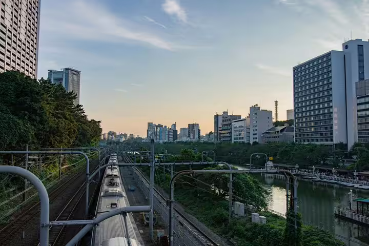Tokyo cityscape with train tracks running alongside a river canal at dusk, surrounded by lush greenery and urban buildings