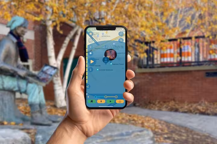 Hand holding smartphone with Tom Thomson mystery tour app near an autumn statue landmark on a self-guided GPS walk.