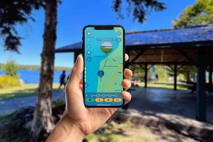 Hand holding smartphone displaying GPS map for Tom Thomson self-guided walking tour near a lakeside park pavilion.