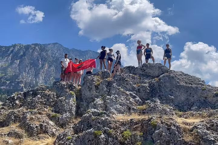 Group of hikers holding a red flag on a rocky summit at Tomor Mountain, enjoying the sunny skies and scenic views of Dardhe.