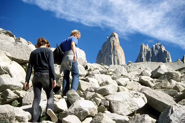 Two trekkers ascend rocky terrain towards the majestic Torres del Paine, set against a vibrant blue sky.