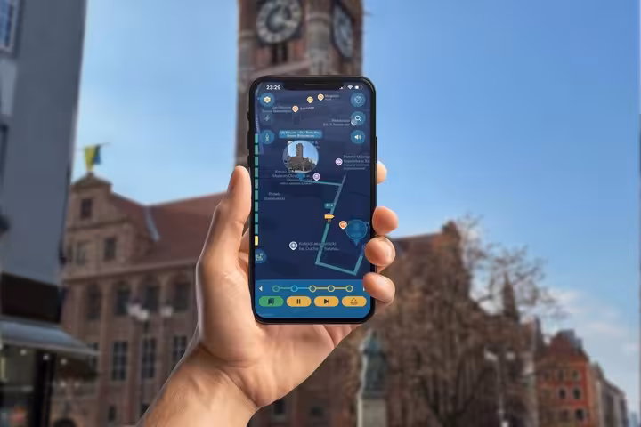 Hand holding smartphone displaying Torun self-guided GPS tour app with historic clock tower in background