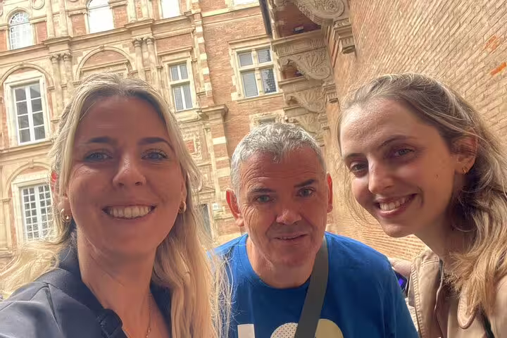 Three smiling tourists posing for a selfie in front of Toulouse's historic pink brick architecture