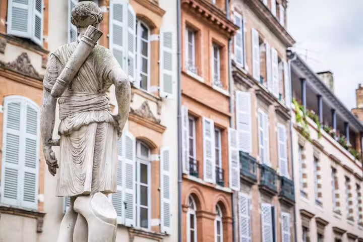 Classical stone statue facing colorful pink buildings with white shutters in Toulouse, France