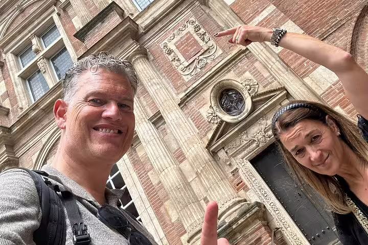 Two tourists smiling in front of a historic pink brick building in Toulouse, France