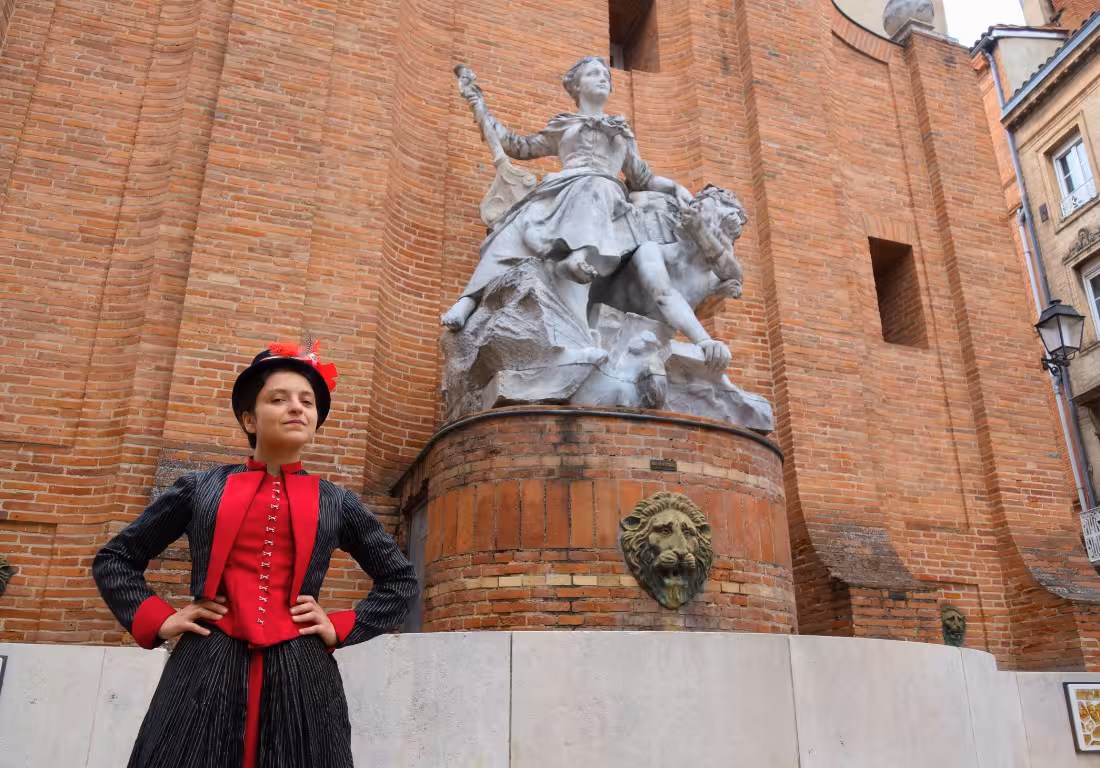 Guide in 19th-century costume stands before a marble statue and brick wall in Toulouse Saint Georges district