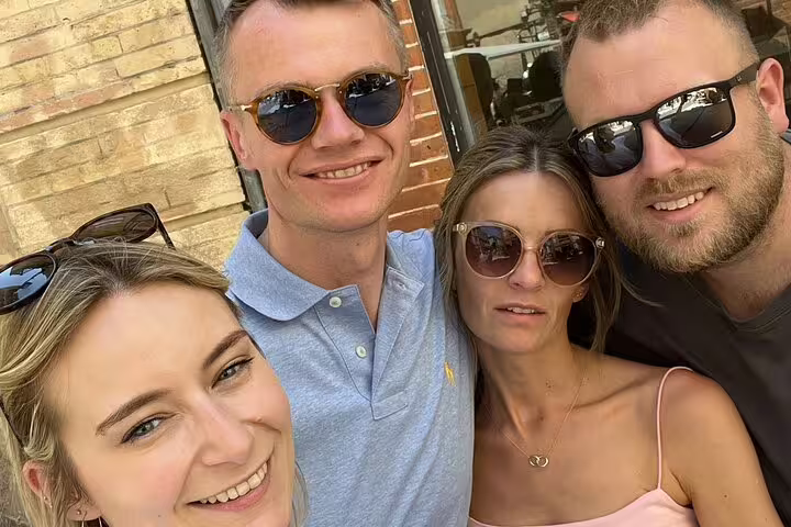 Four smiling friends posing for a selfie outdoors near a brick wall on a sunny day