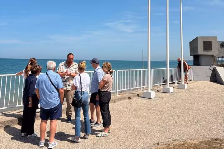 Tour guide speaking to a small group of visitors on a seaside promenade overlooking Omaha Beach, Normandy.
