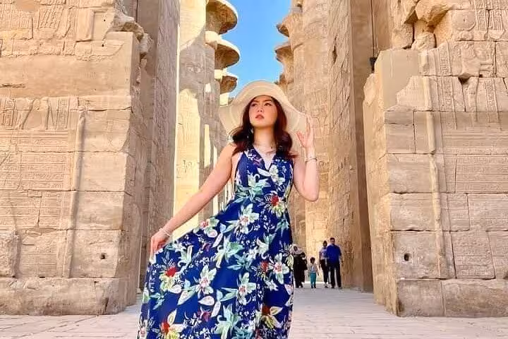 A tourist in a floral dress and sun hat poses amid the towering stone architecture of Karnak Temple.