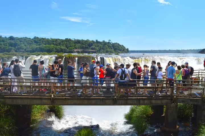 Tourists on a bridge admire the majestic Iguazu Falls under a clear blue sky during a private guided tour.